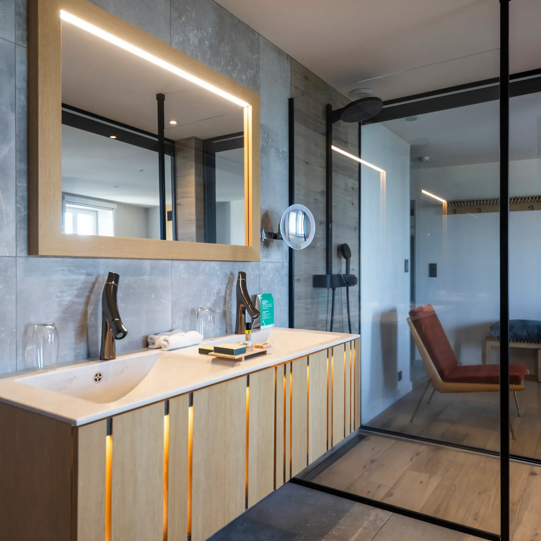 Salle de bain moderne avec lavabo en pierre, miroir éclairé et douche vitrée, vue sur une pièce adjacente avec chaise et banc
