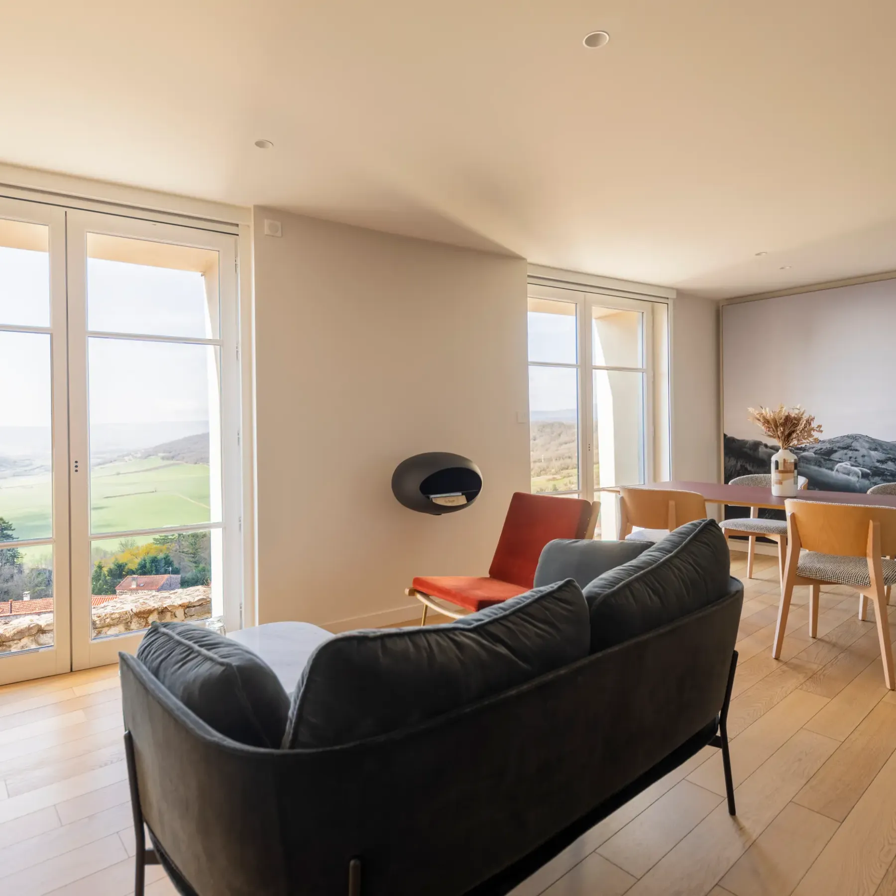 Un salon avec canapé noir et table à manger en bois, donnant sur une vue extérieure par de grandes fenêtres