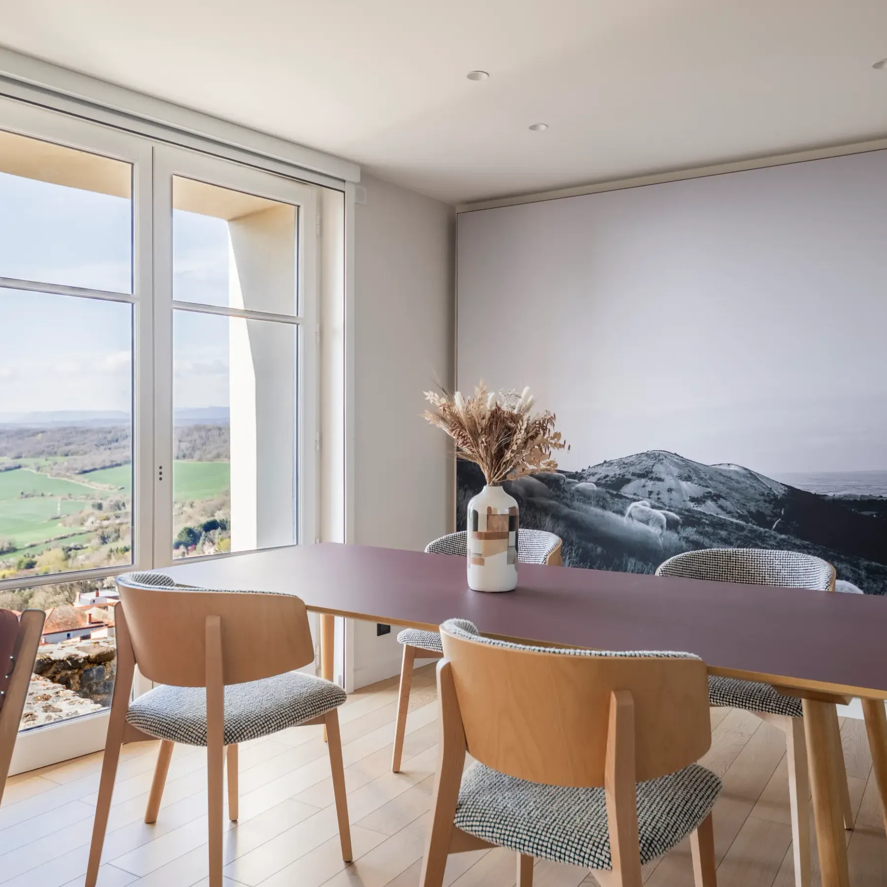 Une table rectangulaire avec des chaises autour, devant une grande fenêtre offrant une vue sur un paysage extérieur