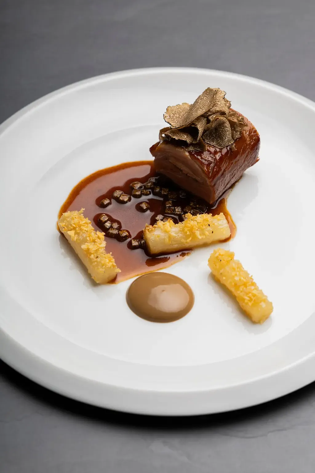 A piece of meat with a slice of truffle, served with sauce and two golden sticks, presented on a white plate.