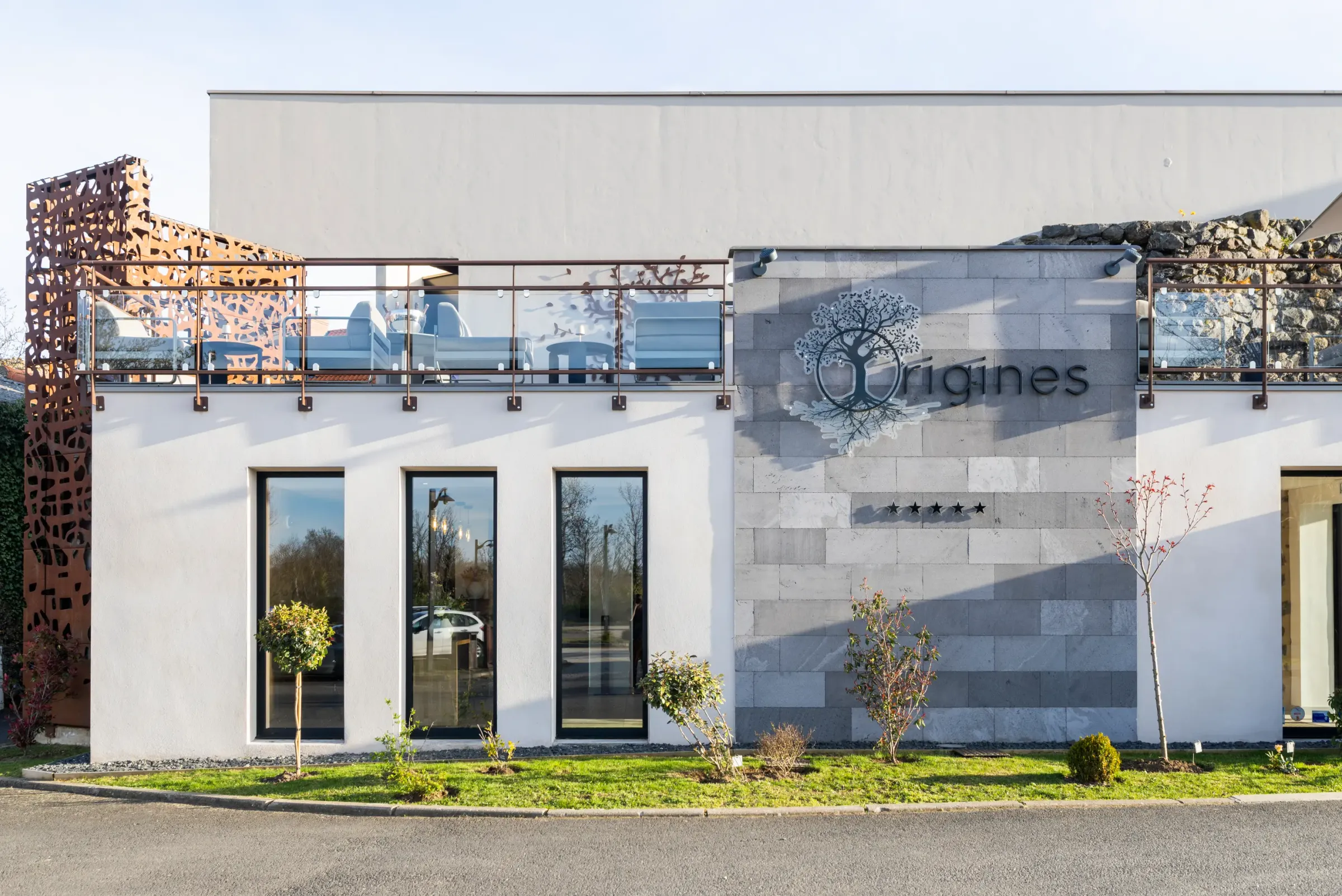 Modern building with white and grey facade, metal and vegetated balcony railing, and "Origines" sign with a tree logo