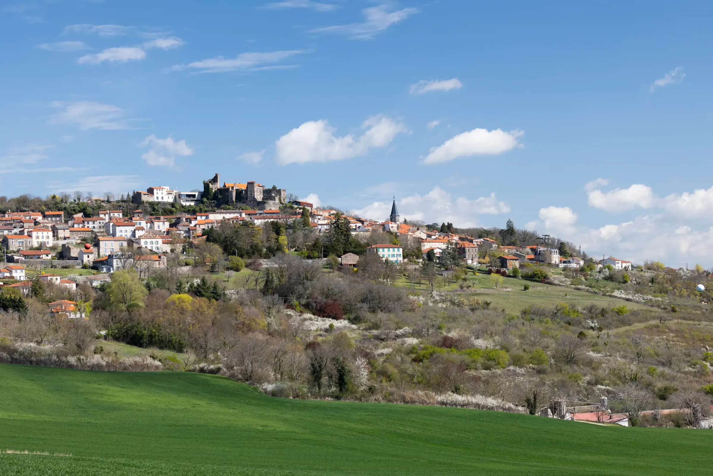 Un village perché sur une colline avec des maisons aux toits rouges, un château en ruine et une église, entouré de verdure et de champs