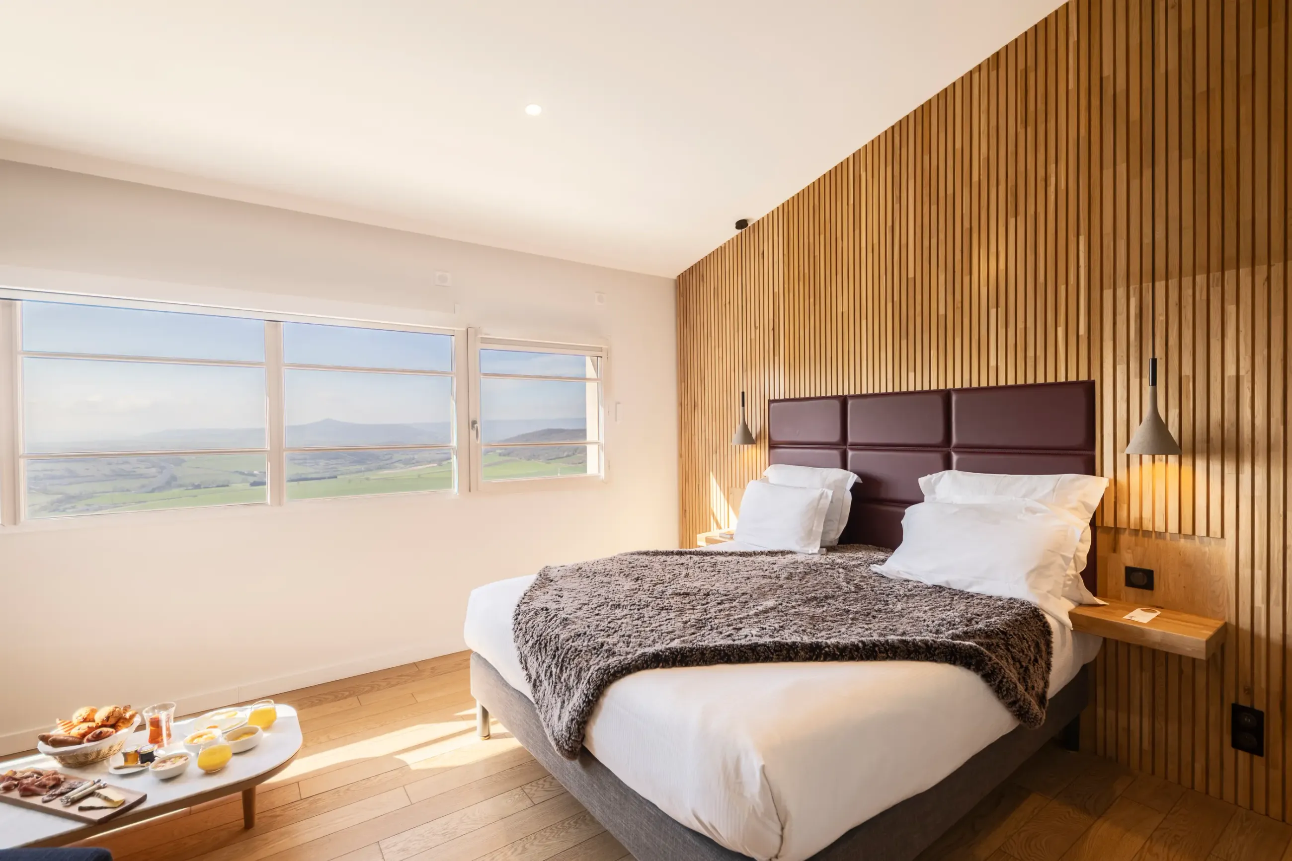 Chambre avec un lit double, une tête de lit en cuir marron, des fenêtres offrant une vue sur un paysage vallonné et une table avec des aliments et des boissons