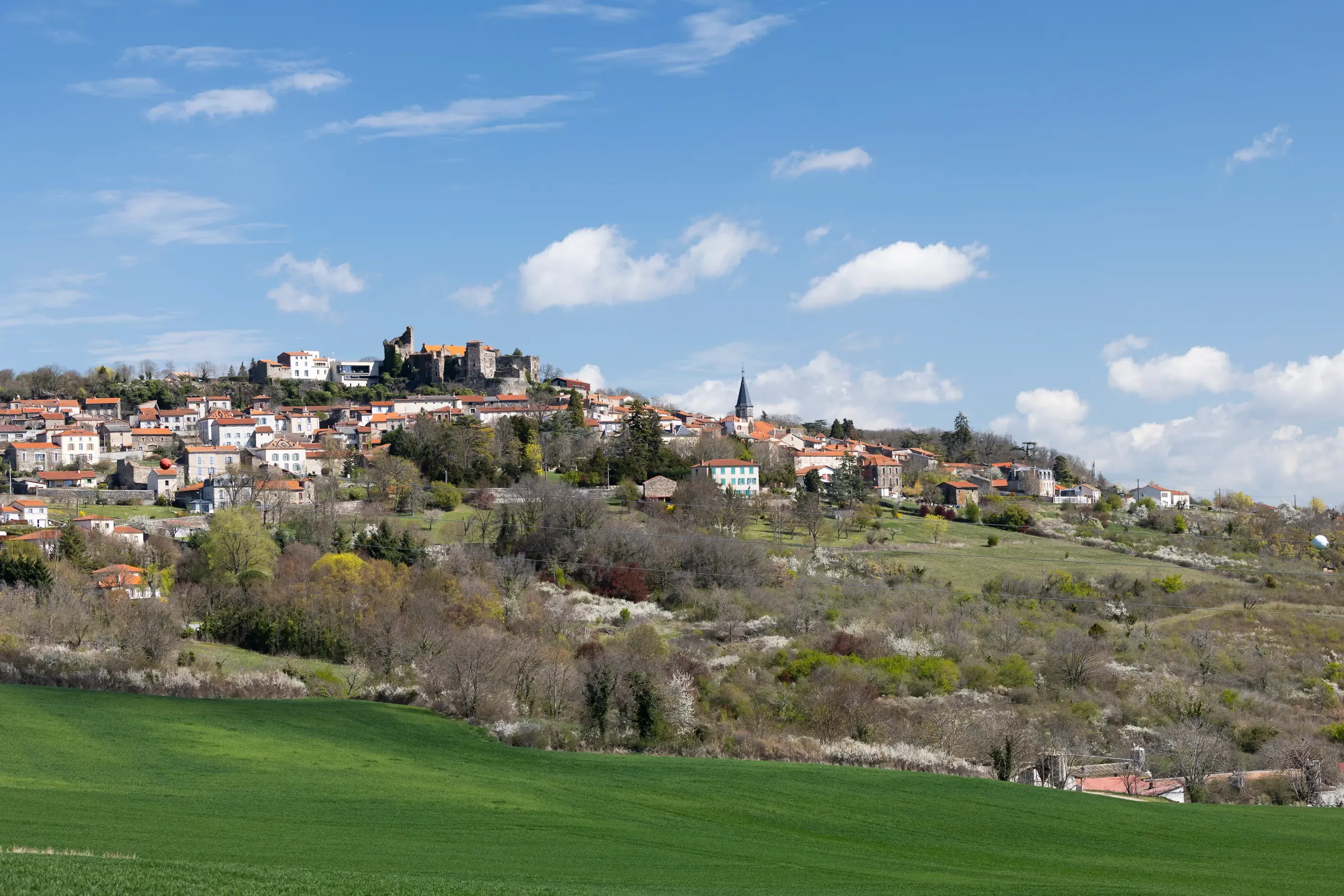 Un village perché sur une colline avec des maisons aux toits rouges, un château en ruine et une église, entouré de verdure et de champs