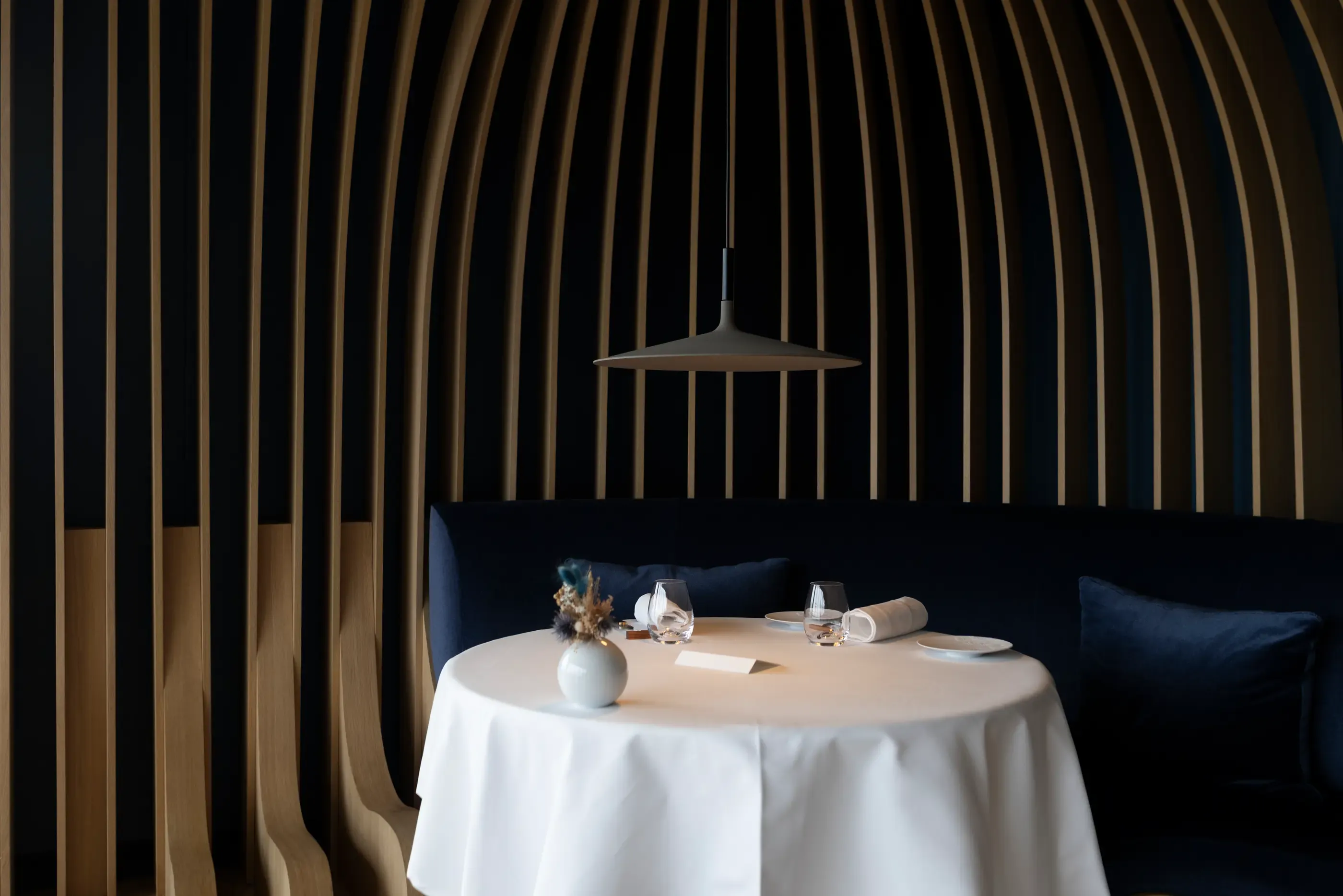 Table set with white tablecloth, flower vase, glasses, and rolled napkin, under a pendant light, in a space with dark and light vertical striped walls.
