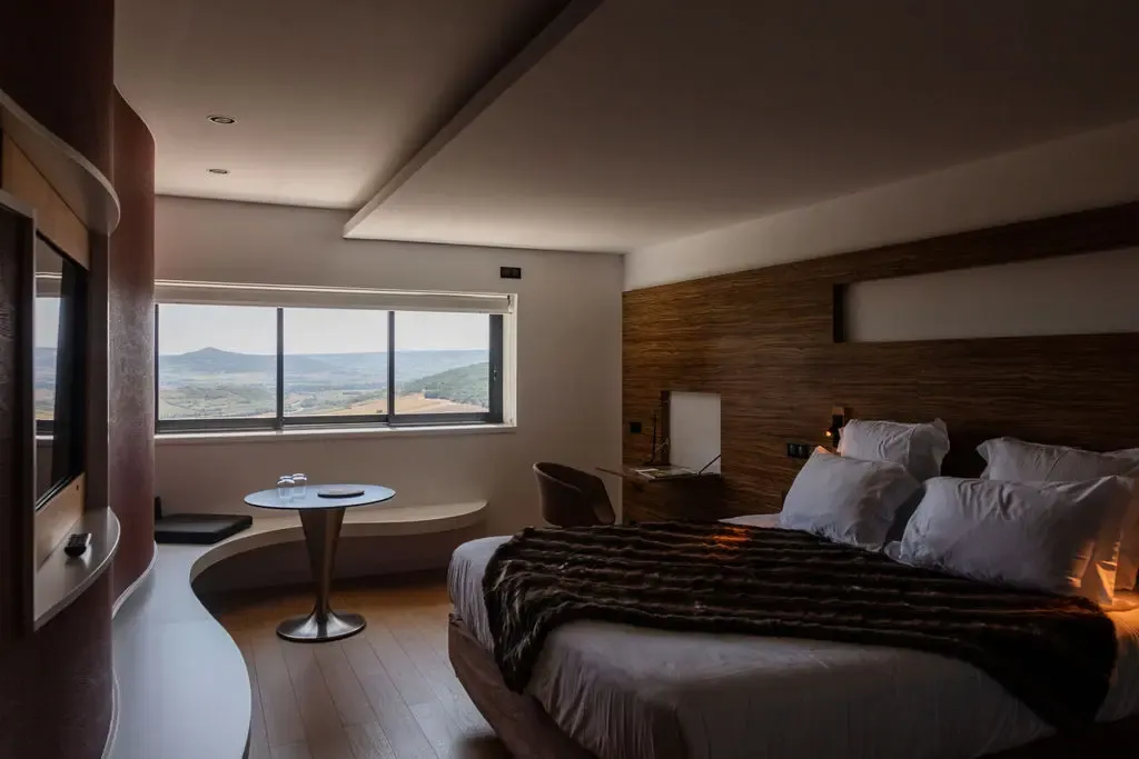 Room with bed, round table and panoramic view of mountains through large windows