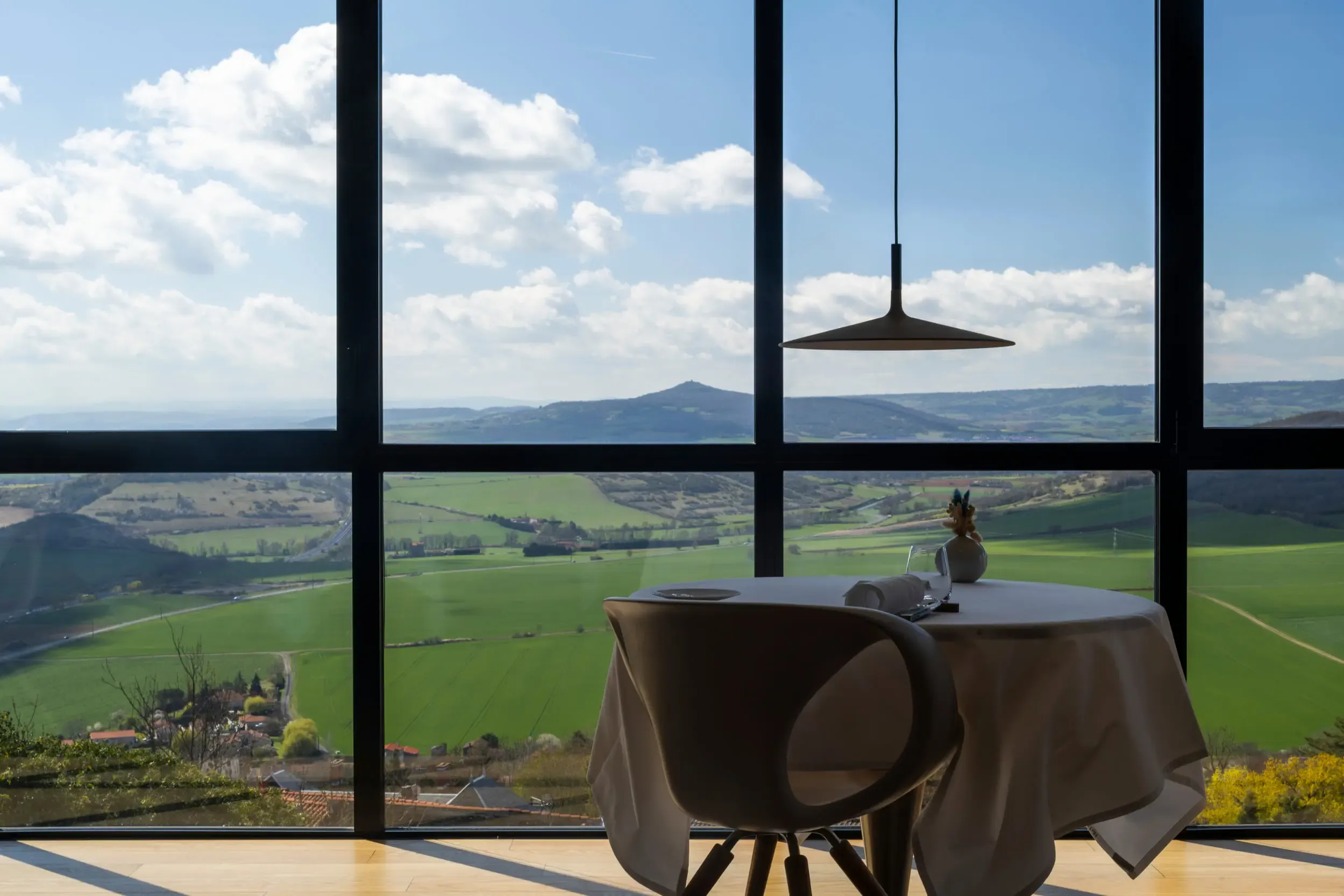 Une table avec une nappe et un vase de fleurs devant une grande fenêtre offrant une vue sur des collines verdoyantes et un ciel partiellement nuageux