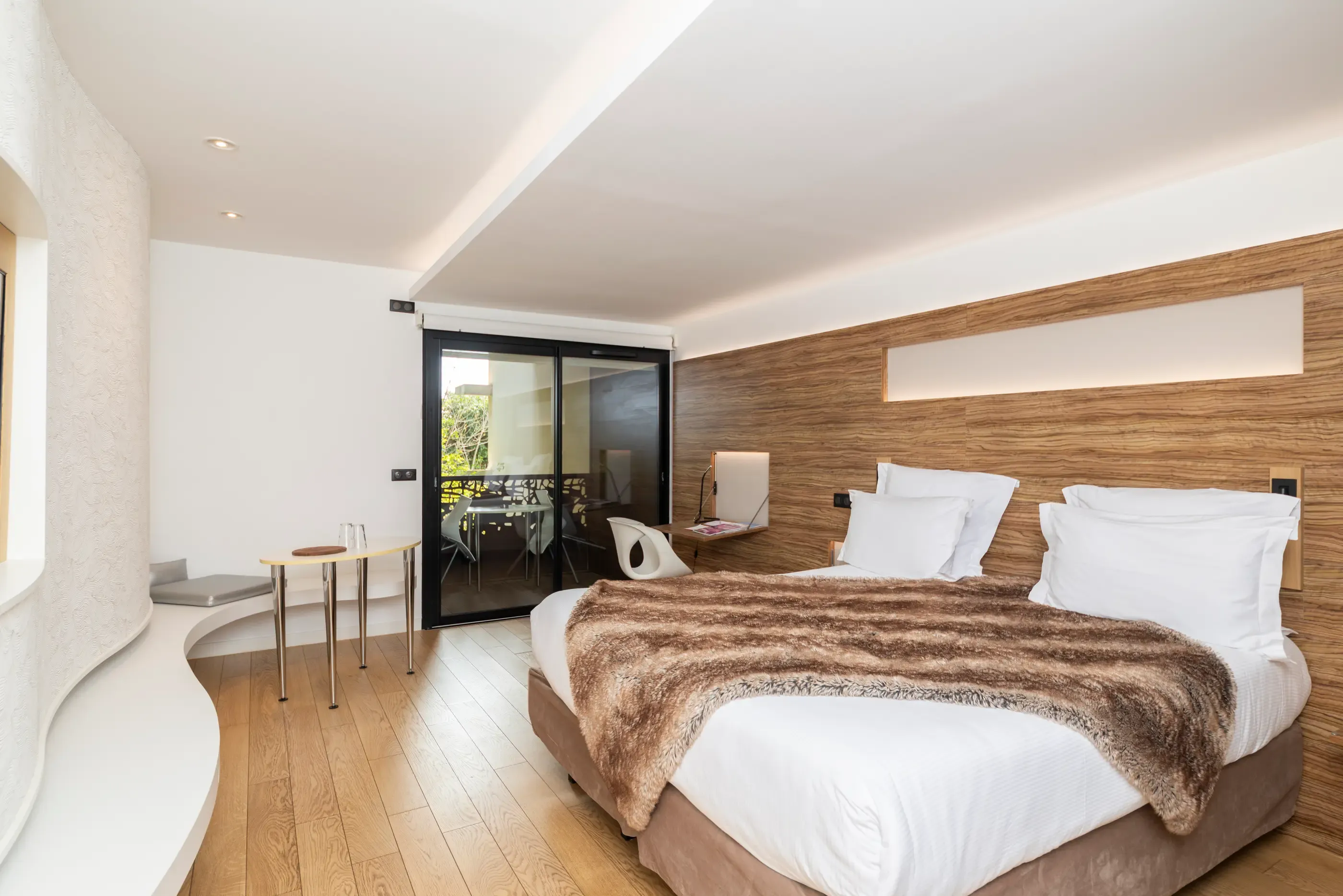 Chambre moderne avec un lit double, une table de chevet, une porte-fenêtre donnant sur un balcon et un mur en bois