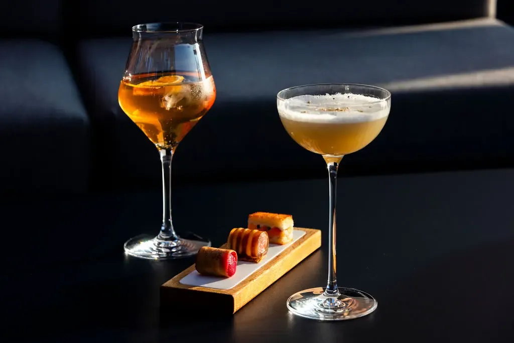 Deux verres contenant des boissons différentes, l'un avec un liquide trouble et l'autre avec un liquide clair, accompagnés de trois petits biscuits sur une planche en bois
