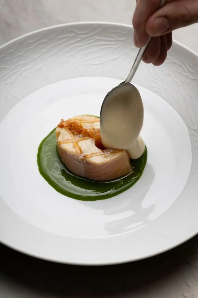A piece of grilled fish is placed on a green sauce, with a white sauce added by the spoonful.
